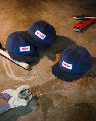 Three blue flatbill caps with labels on a concrete floor with tools in the background