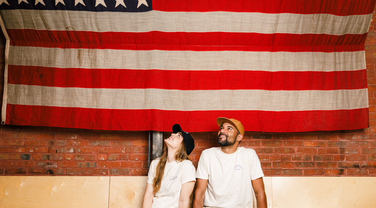 Two people sitting on a couch looking up at a large American flag against a brick wall.